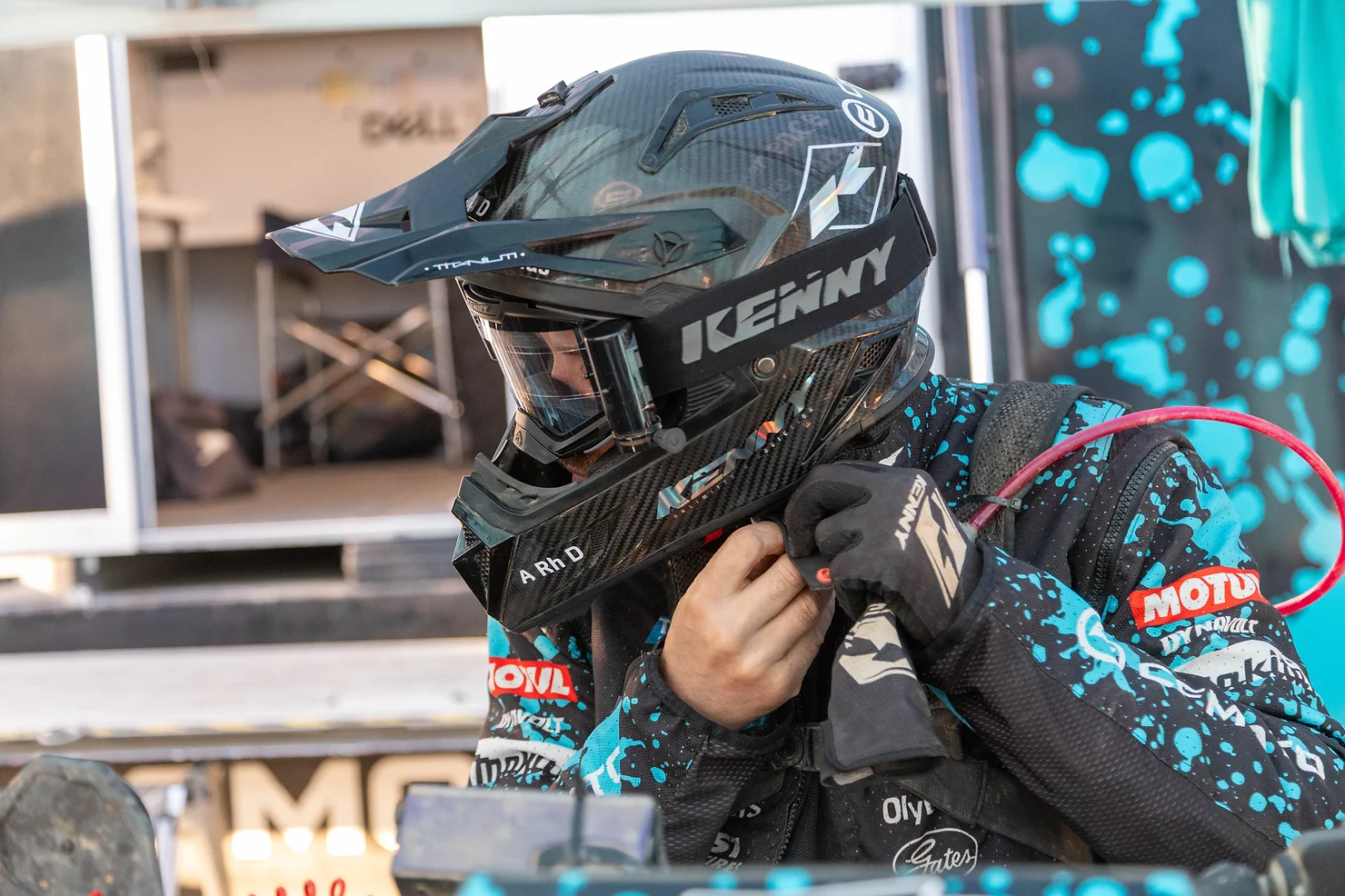 A person wearing a black helmet and a teal-accented racing suit adjusts their gear, ready for the challenge. They stand near a racing vehicle emblazoned with logos and a blue splatter design, representing their dedicated team.