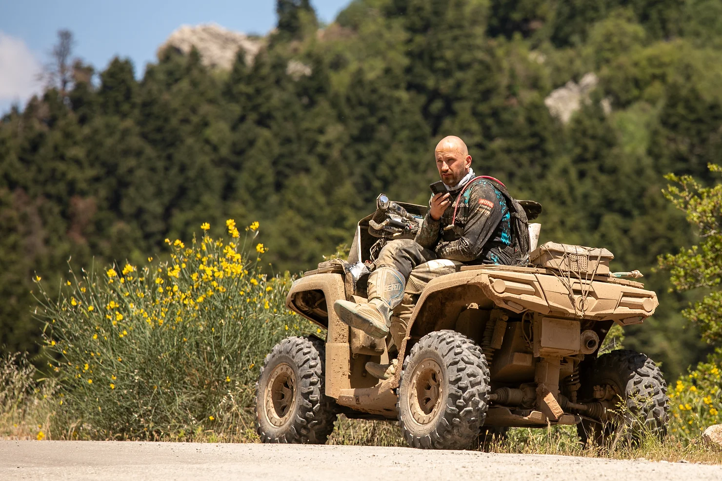 A man sits on a muddy ATV, holding a walkie-talkie amidst the vibrant yellow flowers and trees, prepared for the challenging marathon through Greece's rugged terrain.