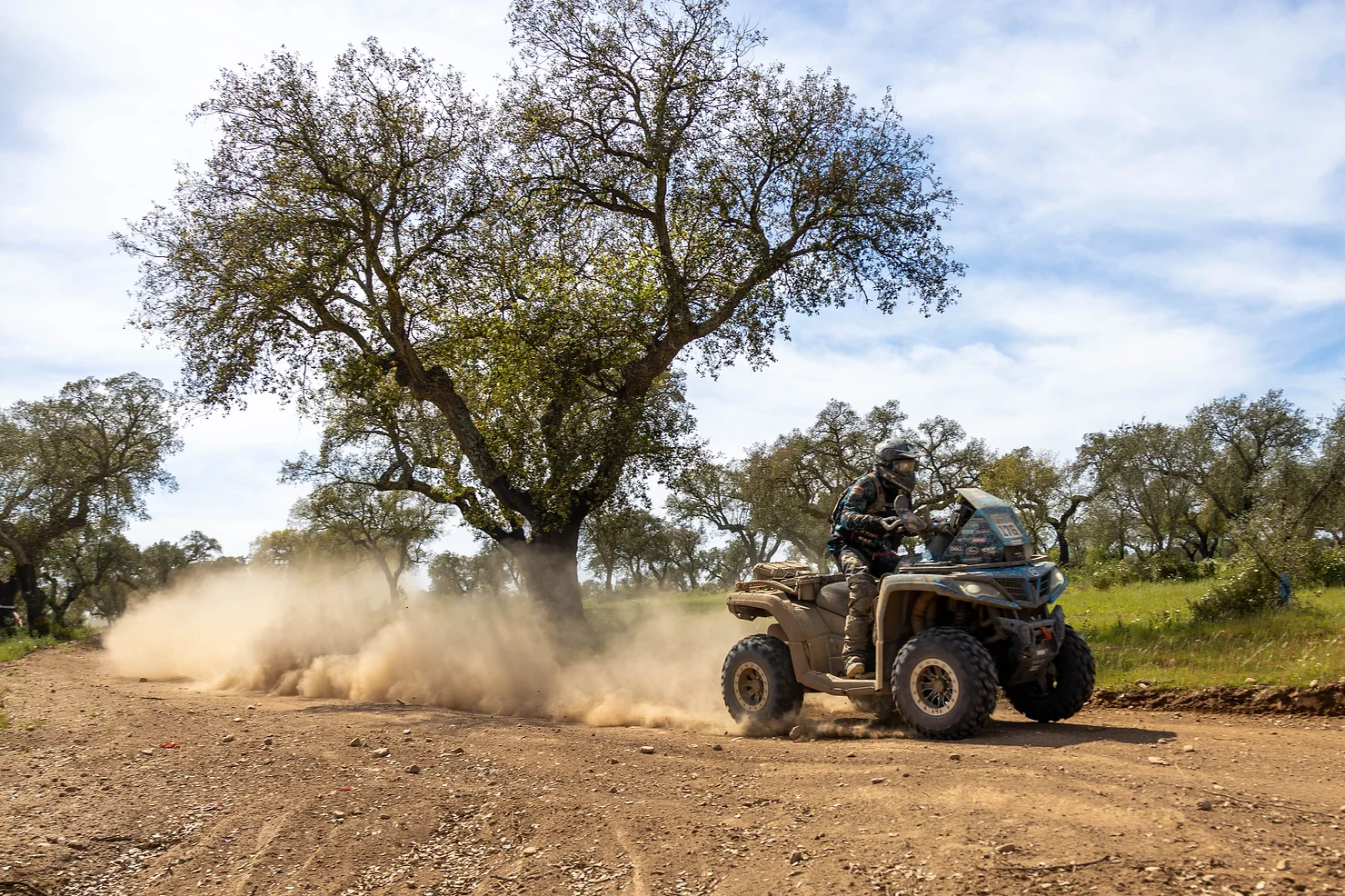 A person is riding an ATV on a dirt path, kicking up a cloud of dust with trees and grass in the background, as if they were performing Sirtaki with the CFMOTO Thunder Racing Team in Greece.
