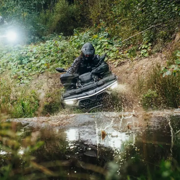 A person expertly maneuvers a quad bike through a muddy water puddle in a forested area, with bright lights shining brilliantly from the vehicle.