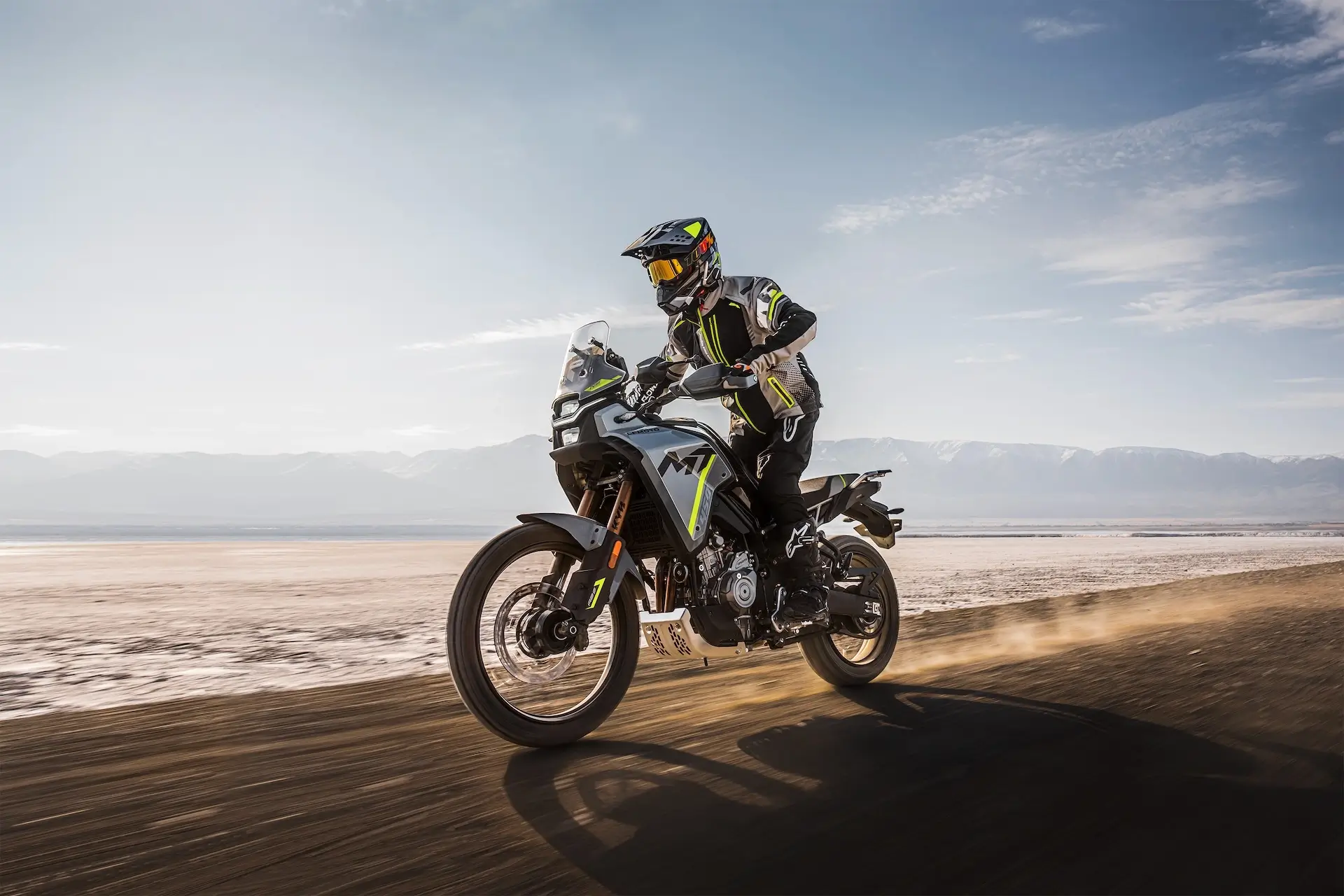 A motorcyclist in full riding gear rides a CFMOTO MOTORCYCLES adventure bike on a dirt road, with mountains and a clear sky in the background.