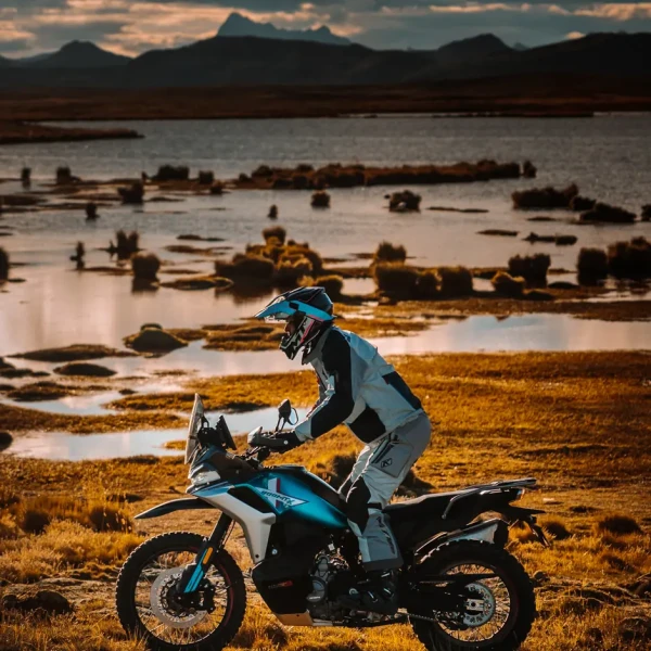 800mt-x feature 3 A person wearing motorcycle gear rides an 800MT-X adventure motorcycle across a grassy landscape near a body of water at sunset.
