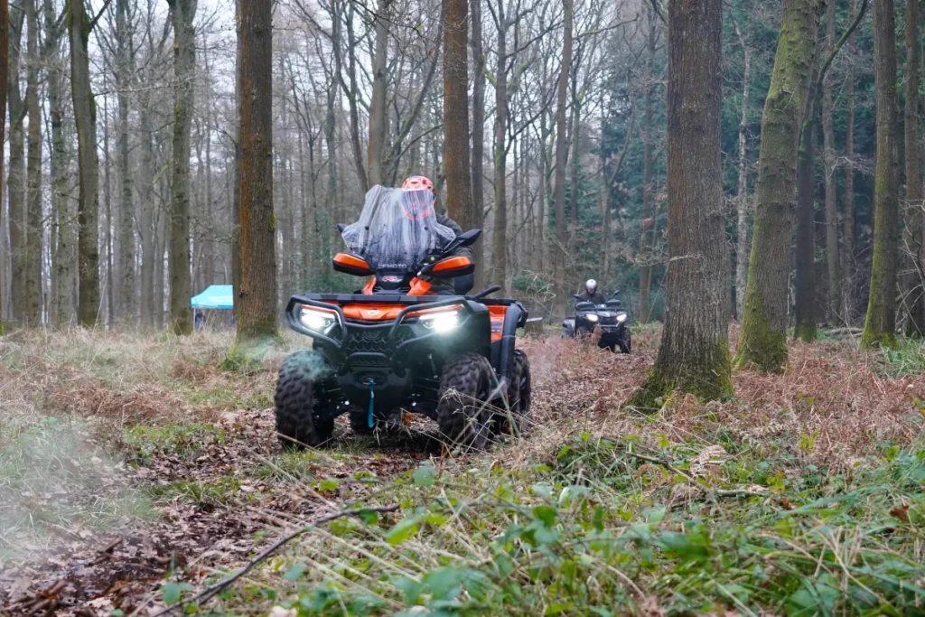 CFMOTO CFORCE 850 TOURING 2 Two people ride CForce 850 Touring all-terrain vehicles along a dirt path through a forest with bare trees and scattered foliage on the ground.