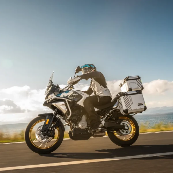 700mt feature 5 A person rides a white 700MT adventure motorcycle with storage boxes on a paved road near a body of water under a partly cloudy sky.