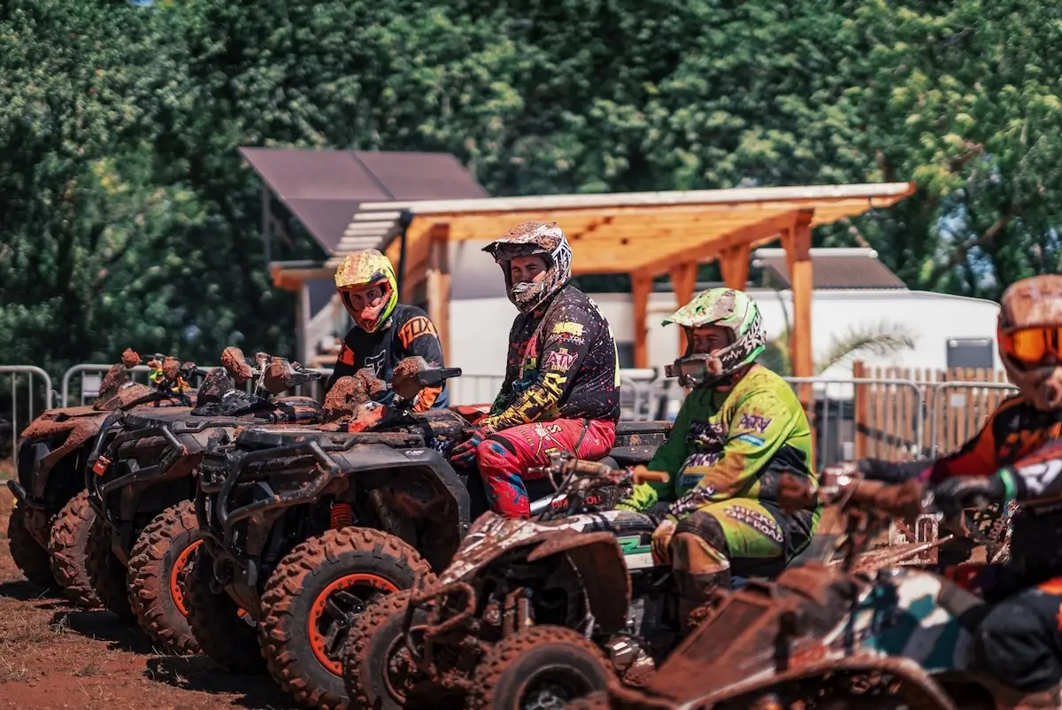 Four people wearing helmets and racing gear sit on muddy CFMOTO CFORCE 1000 ATVs, lined up and waiting on a dirt track with trees and a wooden structure in the background, ready to compete for a podium finish in the 4x4 class.
