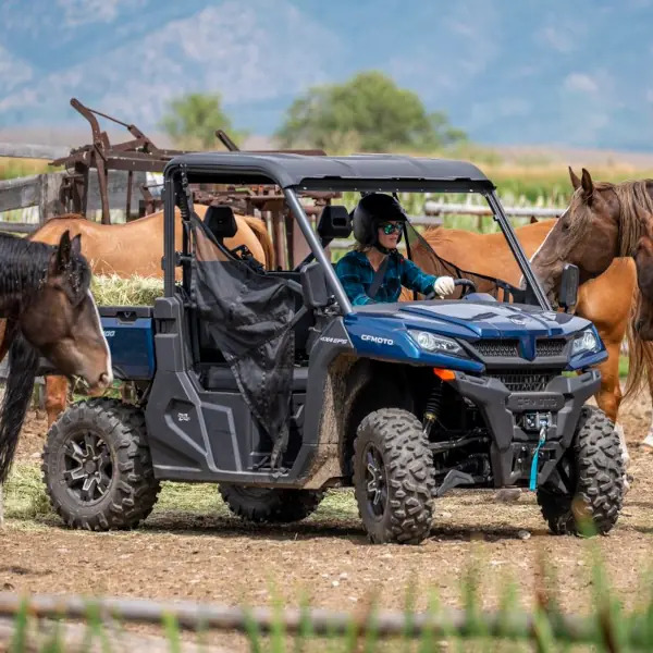 cfmoto horse yard utv A person wearing a helmet drives a blue side-by-side utility vehicle near several equestrian horses in a fenced outdoor area.