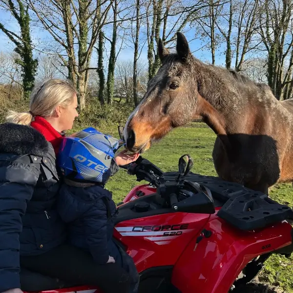 equestrian atv A woman and a child on a red ATV feed a brown horse in a grassy, tree-lined area on a sunny day, enjoying an equestrian moment together.