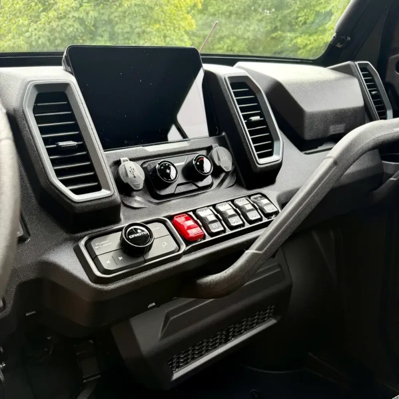 CFMOTO U10 Pro Highland dash Dashboard of the UFORCE U10 PRO HIGHLAND with a central display, control knobs, buttons, and switches, viewed from the passenger side, with trees visible through the windshield.