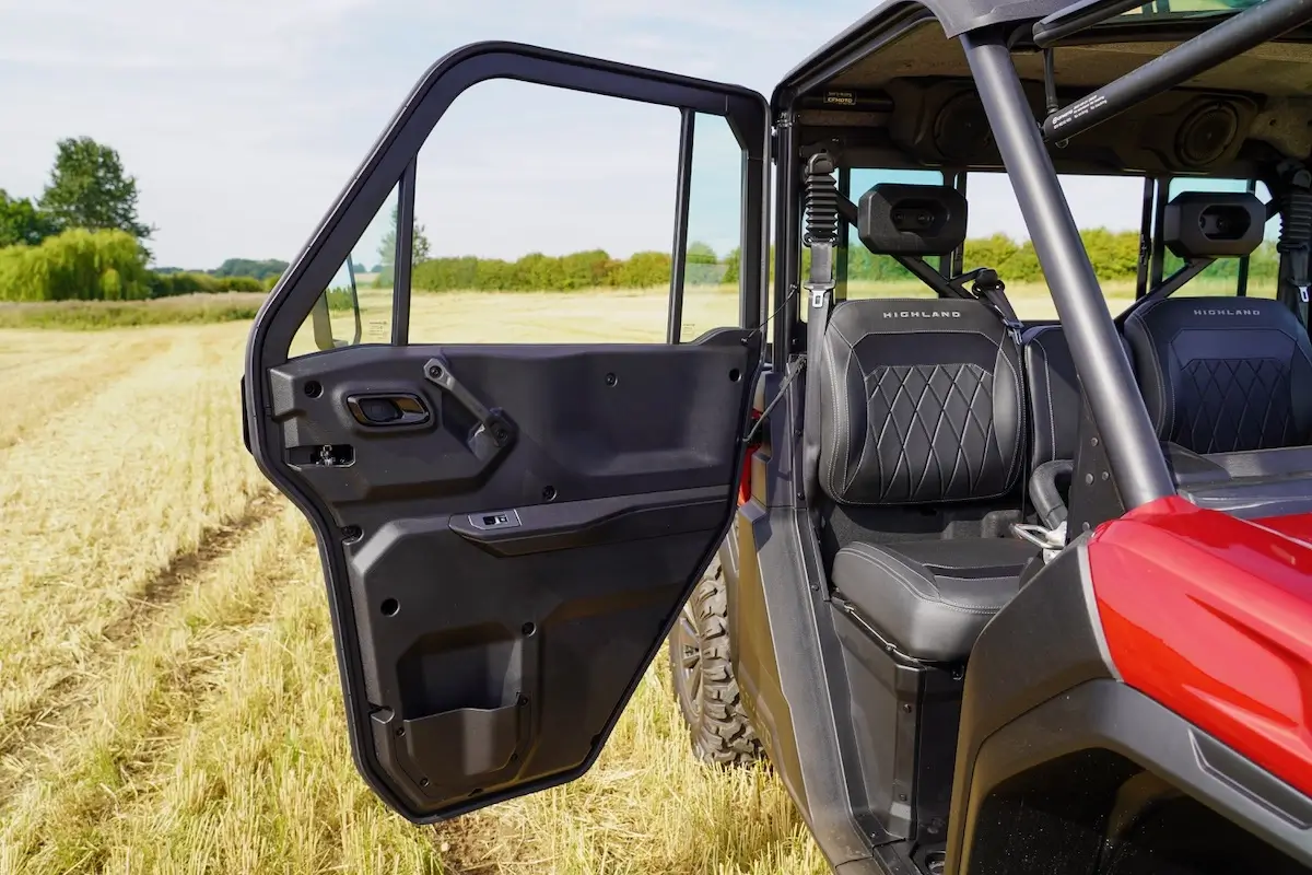 A close-up of an open driver-side door on a red UFORCE U10 PRO HIGHLAND off-road vehicle parked in a harvested field, showing two black seats inside.