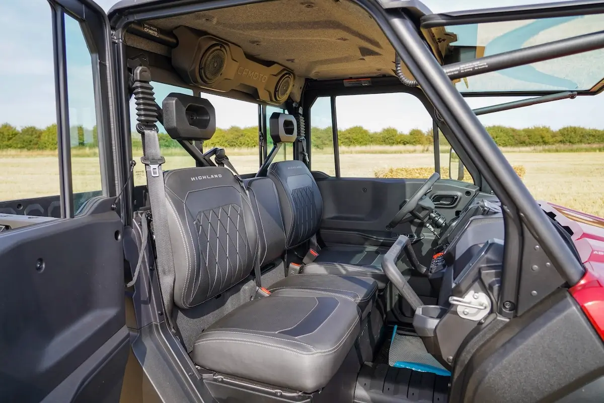 Interior view of the UFORCE U10 PRO HIGHLAND utility vehicle showcasing two front black seats, steering wheel, dashboard controls, and roll bar, with a field and trees visible outside.