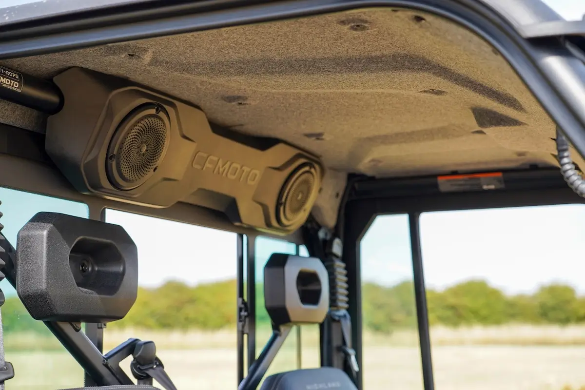 Close-up view of a UFORCE U10 PRO HIGHLAND’s interior roof showing a CFMOTO-branded overhead audio system and headrests, with a window view of trees in the background.