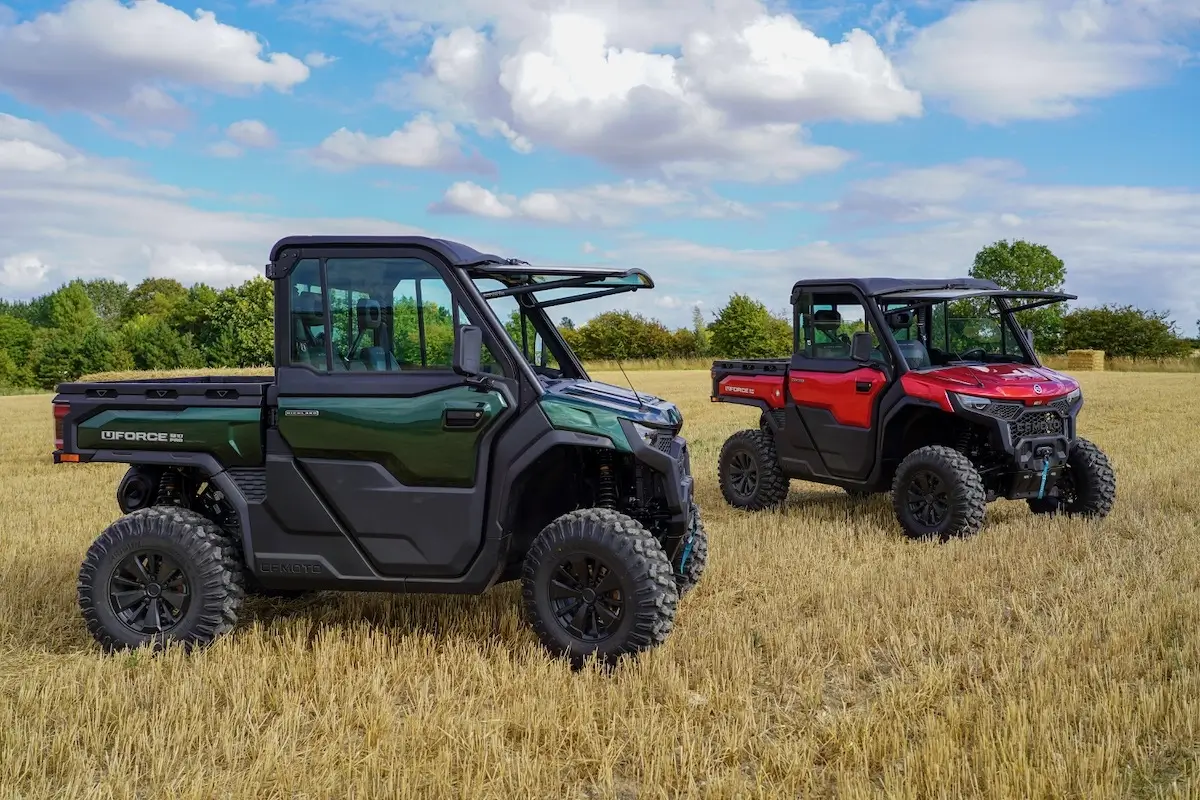Two off-road utility vehicles, one green and one red UFORCE U10 PRO HIGHLAND, are parked on a harvested field under a partly cloudy sky with trees in the background.