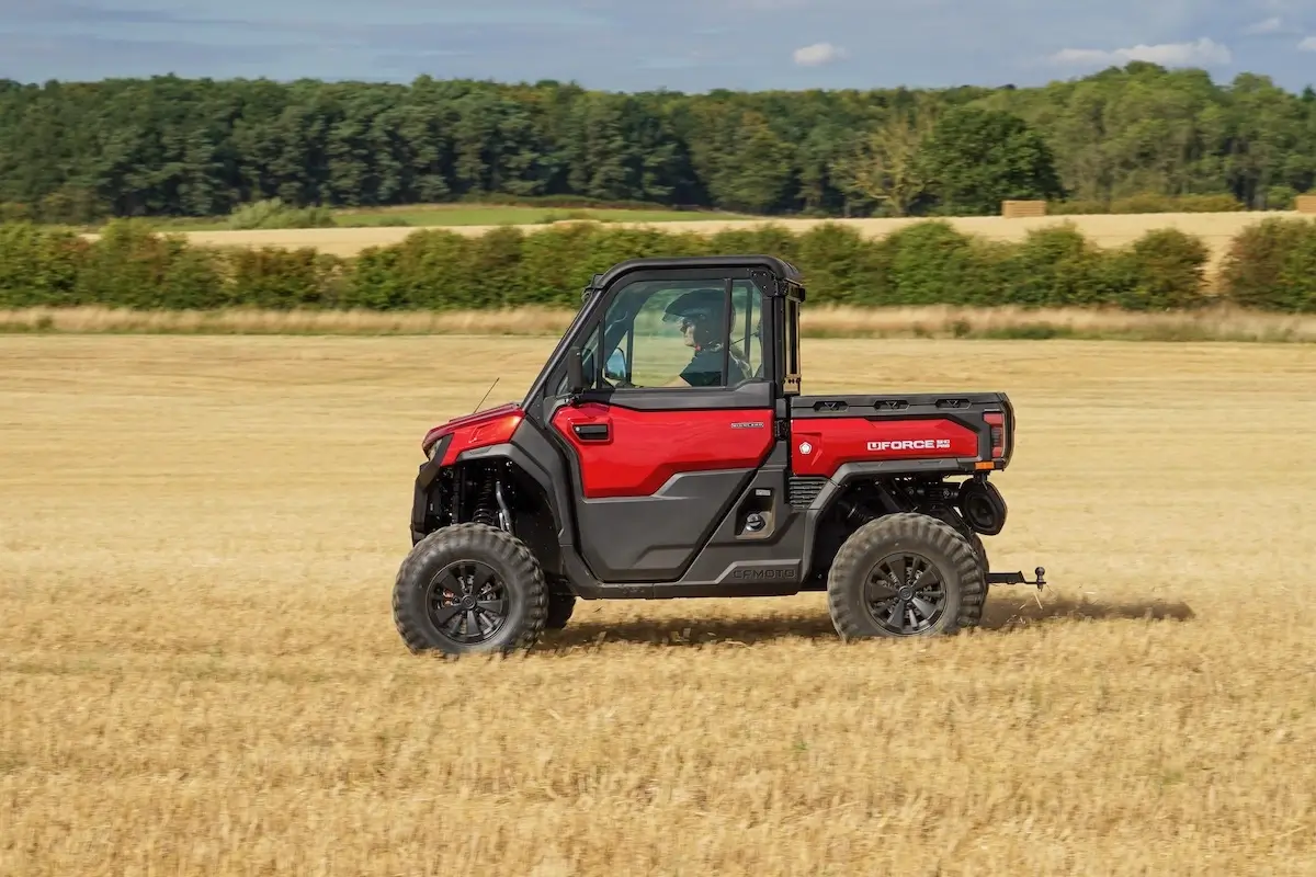 A red UFORCE U10 PRO HIGHLAND utility task vehicle drives through a harvested field, with trees and hedges visible in the background under a partly cloudy sky.