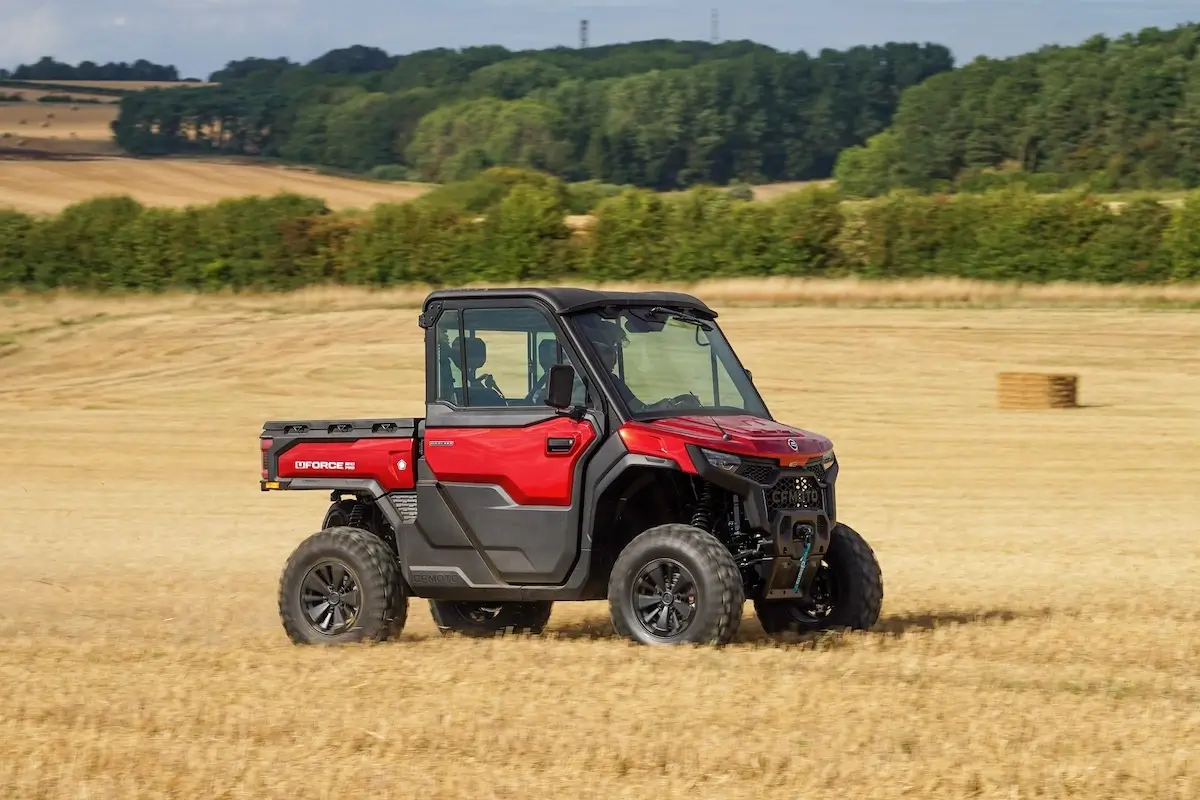 A red UFORCE U10 PRO HIGHLAND utility terrain vehicle with a cab and cargo bed drives across a harvested field, with trees and bales of hay in the background.