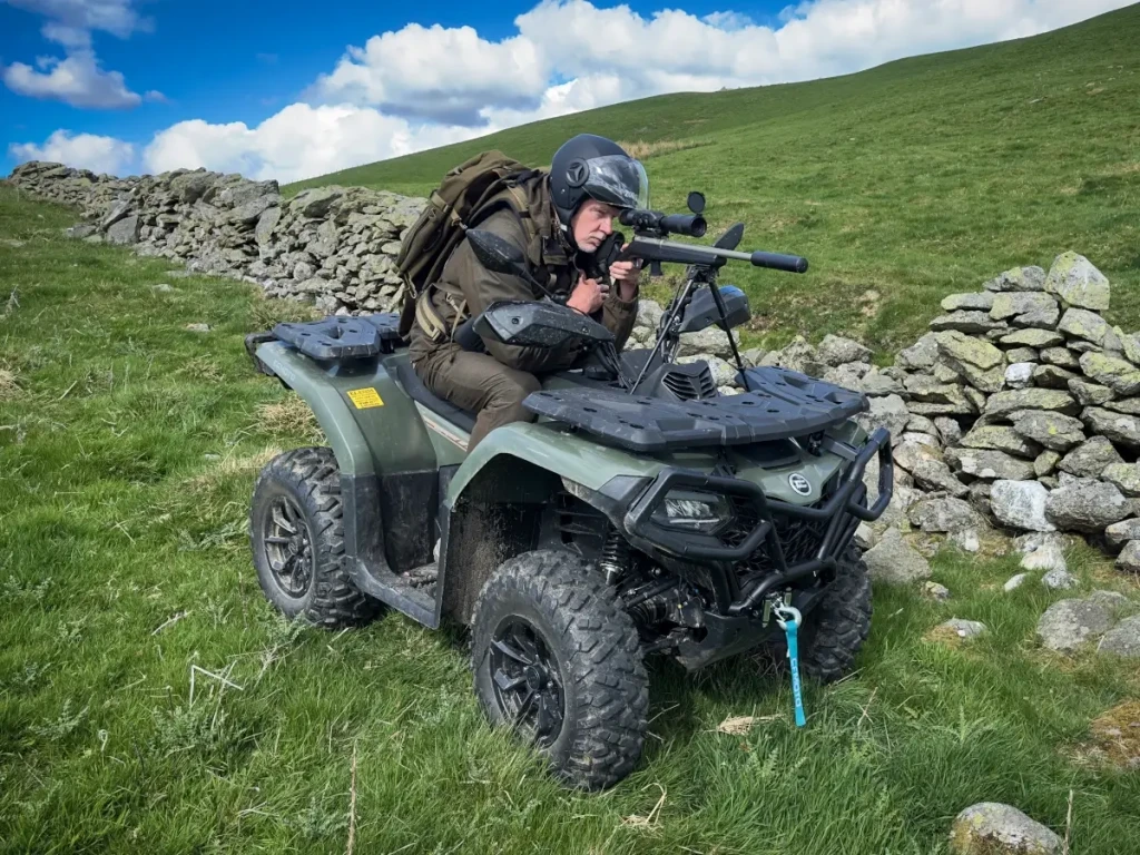 Shooting ATV A person wearing a helmet and outdoor gear, engaged in estate management, aims a rifle while seated on an all-terrain vehicle in a grassy, rural landscape next to a stone wall.