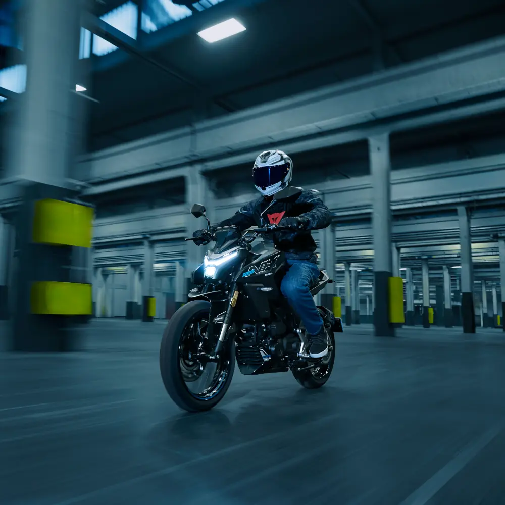 commuter motorcycle 4 A person wearing a helmet and jacket rides a commuter motorcycle through a spacious, industrial-looking indoor area with columns.