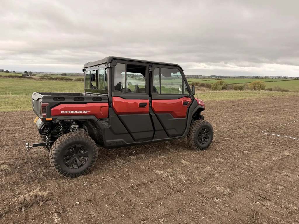 CFMOTO UFORCE U10 XL A red and black UFORCE U10 PRO XL utility vehicle is parked on a dirt field under a cloudy sky, with open farmland stretching out in the background.