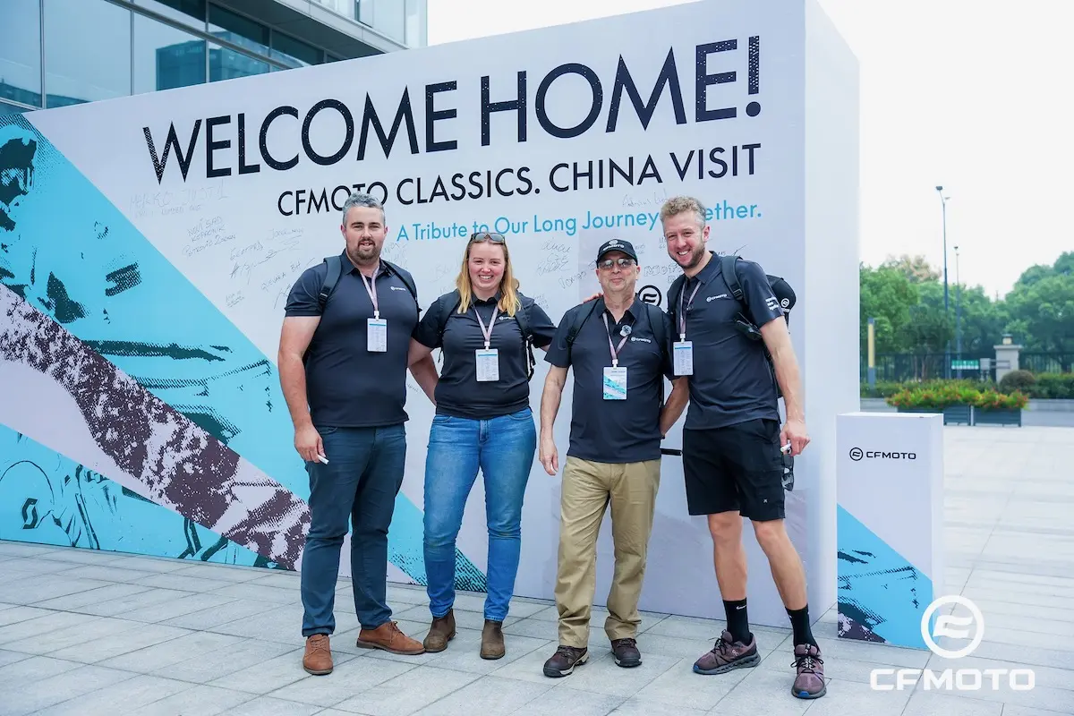 Four people in matching shirts pose in front of a large "Welcome Home! CFMOTO Classics. China Visit" sign outside a modern building, highlighting their shared passion for ATV safety.