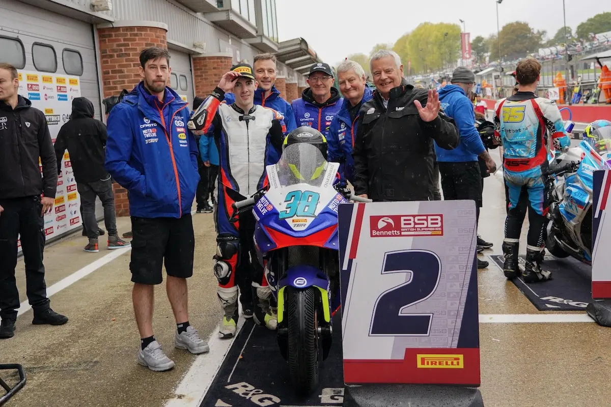 A group of people stand behind a CFMOTO motorcycle marked with the number 38, next to a sign displaying "2" at a Moto Twelve racing event.