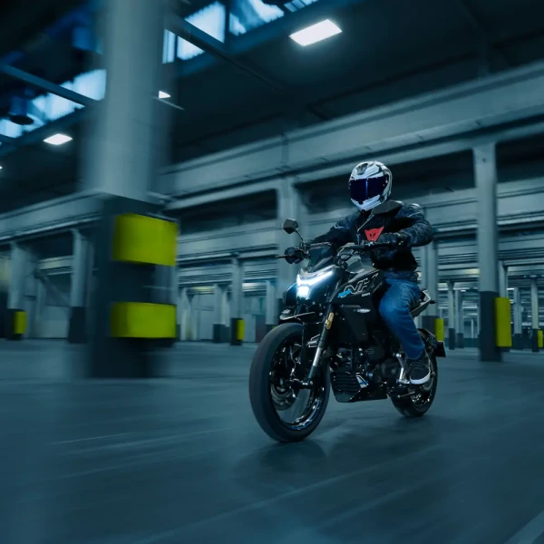 125NK_Gem Black_Action (3) A person wearing a helmet rides a black motorcycle through a large, industrial indoor space with concrete pillars, making it an ideal setting for new riders to practice their skills.