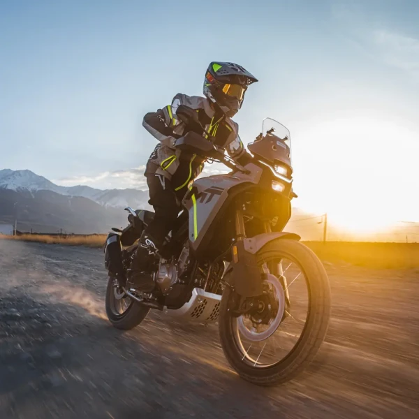 450MT_Outdoor pictures_Others (10) A person in full protective gear rides a motorcycle at speed on a gravel road with mountains and the sun in the background, showcasing an ideal adventure for new riders seeking excitement and skill-building.