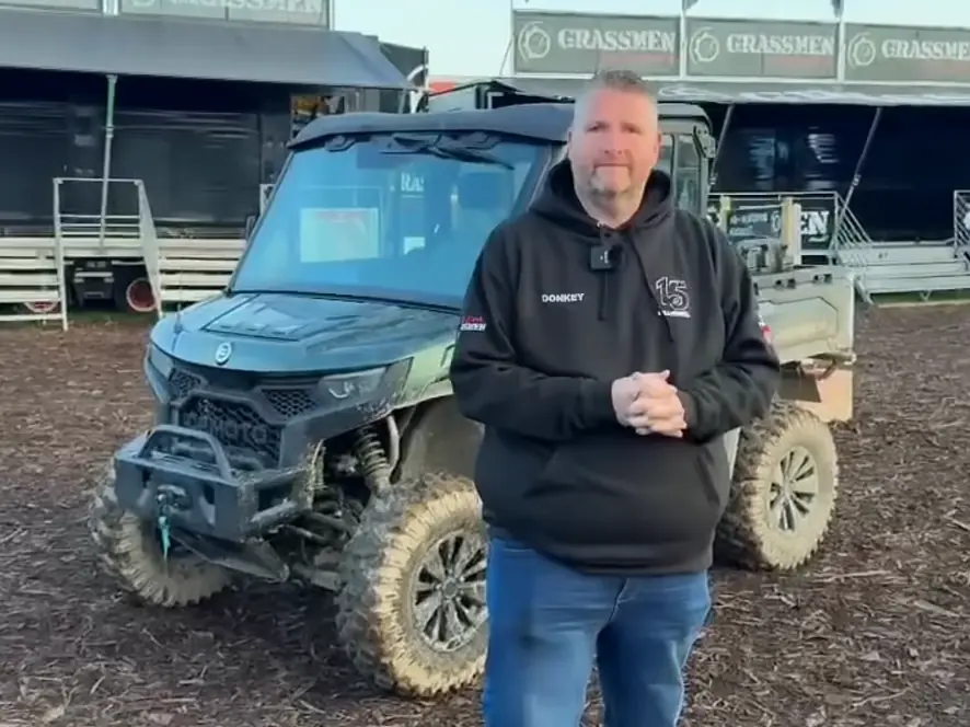 Gareth Donkey from Grassmen and his CFMOTO UFORCE U10 PRO 2 A man wearing a black hoodie stands in front of a muddy CFMOTO CFORCE TECH GEN 4 Concept off-road utility vehicle at an outdoor event with "GRASSMEN" signs in the background.