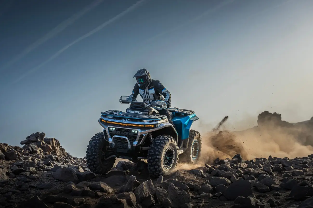 A person wearing protective gear rides a blue CFMOTO classics all-terrain vehicle over rocky terrain, kicking up dust under a clear sky.