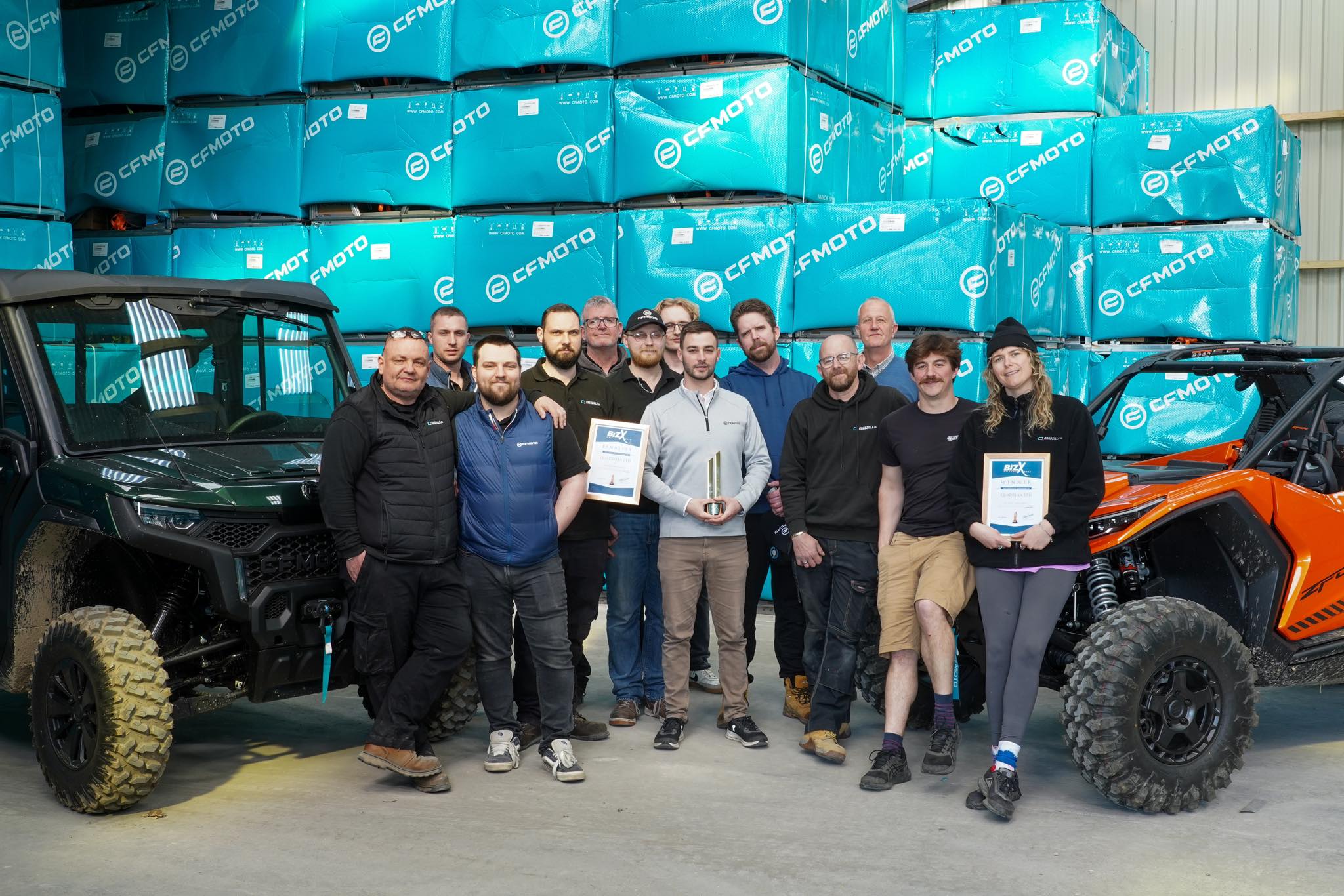 A group of people stand indoors between two off-road vehicles, holding certificates and a trophy, with stacked CFMOTO boxes in the background—highlighting quality despite common concerns about problems with Chinese ATVs.