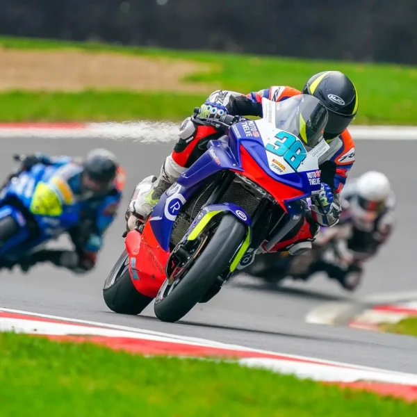 Three racing motorcycles lean into a curve on a track day, with the leading rider wearing number 38 and spraying liquid from the back of the bike.