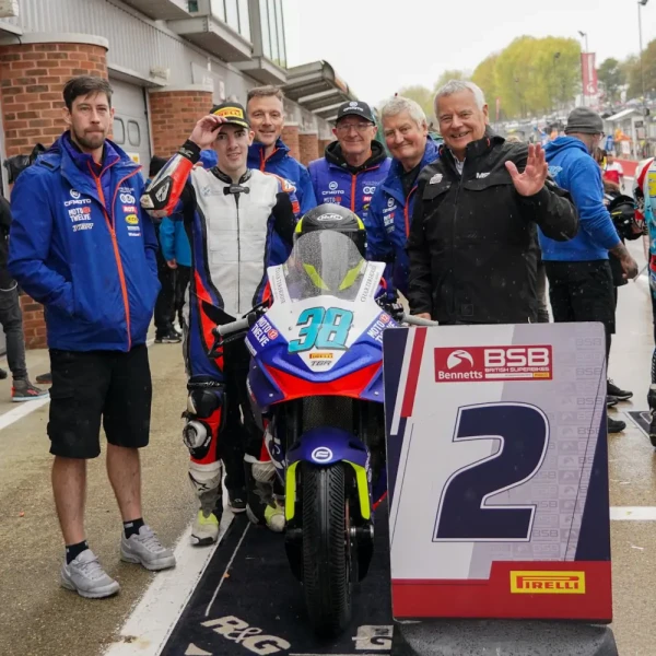 Six men pose next to a racing motorcycle and a sign showing the number 2 at a racetrack event. One man is in racing gear, while the others wear team uniforms, capturing the spirit of an exciting track day.
