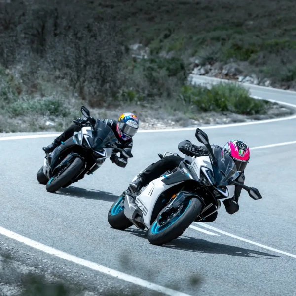 Two motorcyclists wearing helmets ride sport bikes, leaning into a curve on a paved road surrounded by greenery, enjoying the thrill of leisure riding.