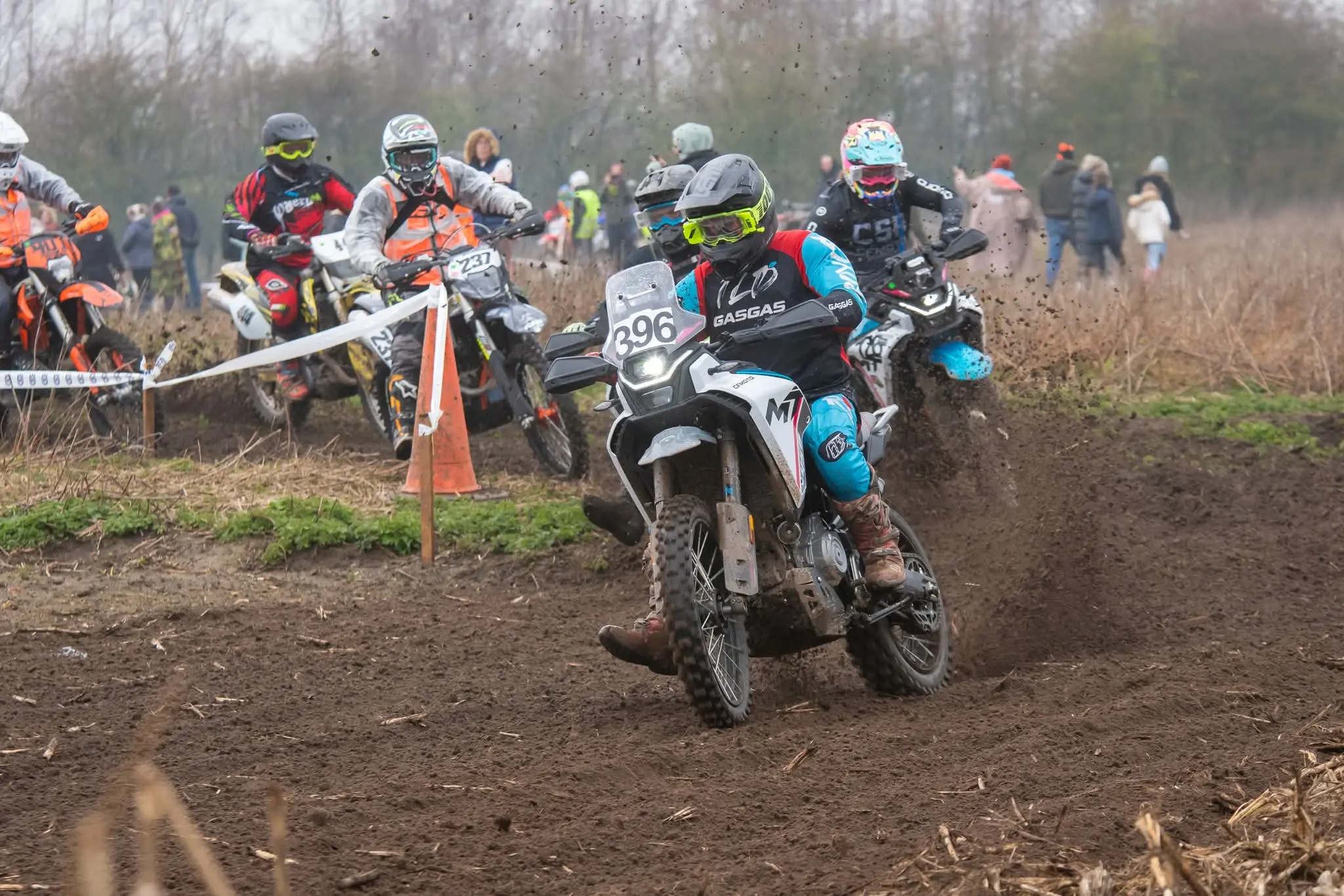 Motorcyclists race on a dirt track at a Lincs Enduro Club event, kicking up mud as they navigate a bend; spectators and officials are visible in the background.