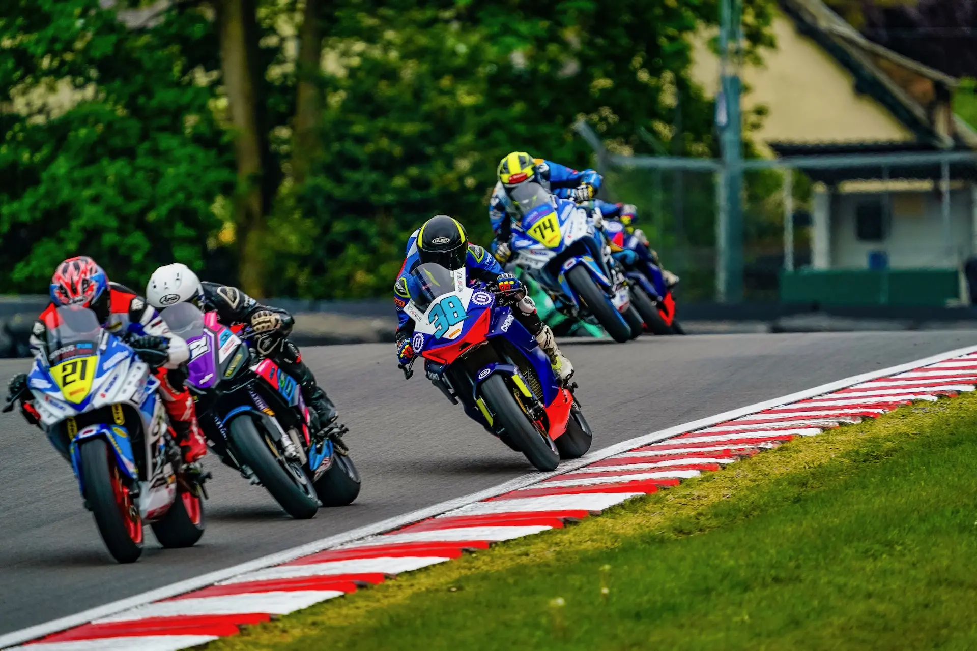 Four motorcycle racers from Moto Twelve lean into a bend at Oulton Park racetrack, closely following each other, with trees and buildings in the background.