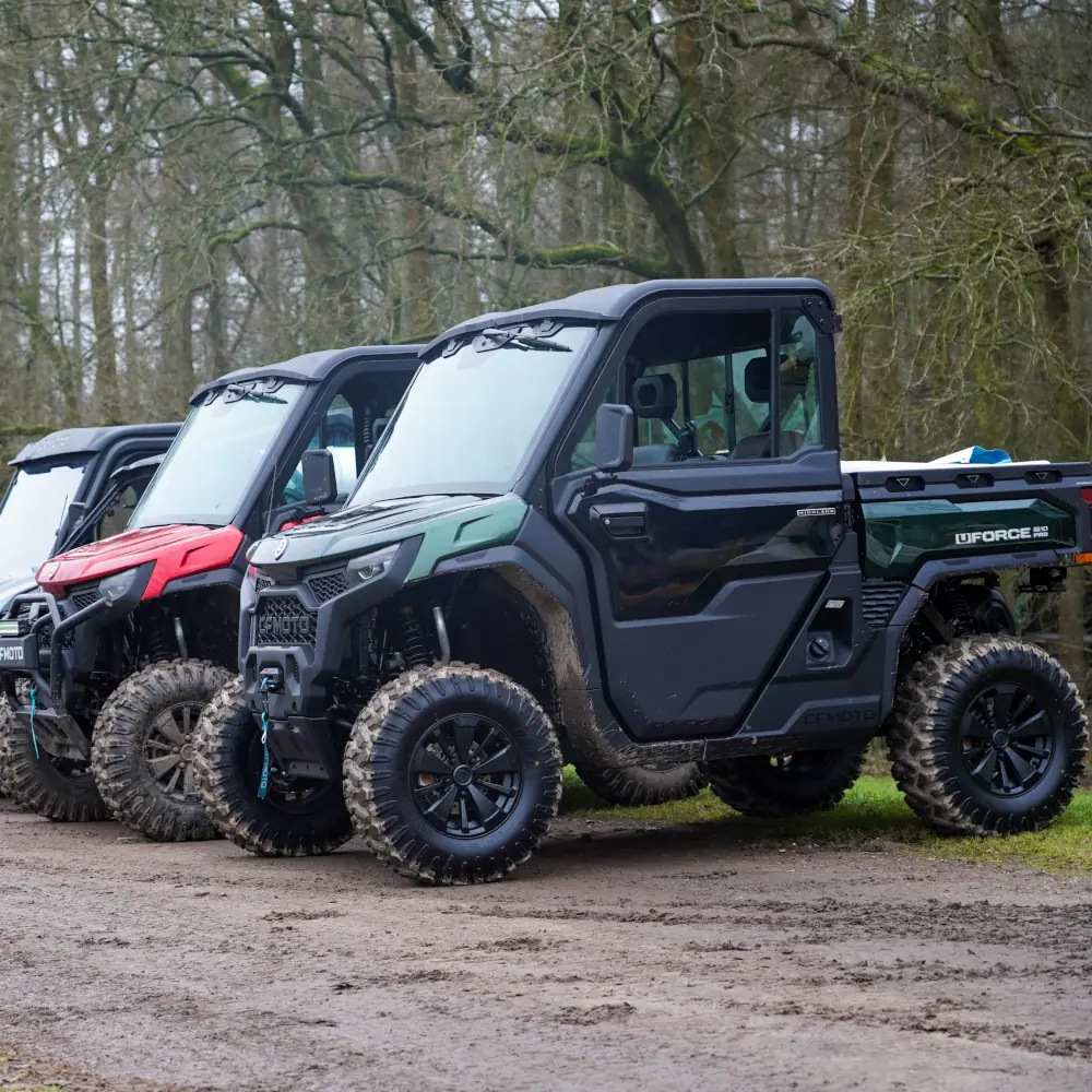 Fleet vehicles A fleet of three off-road utility vehicles with large, rugged tyres is parked on a dirt path beside a wooded area.