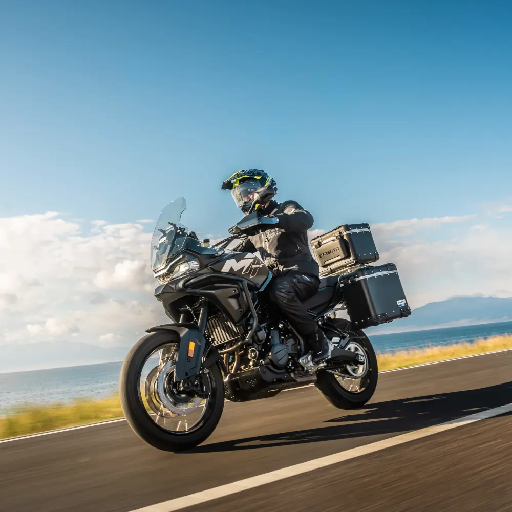fleet motorcycles A motorcyclist wearing kit rides a black touring motorbike from a fleet, with panniers, on a coastal road under a clear blue sky.