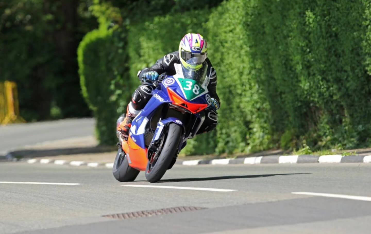 A motorcyclist in racing gear rides a blue and orange CFMOTO sportbike with the number 38 on a road surrounded by greenery, evoking the thrill of the Isle of Man TT.