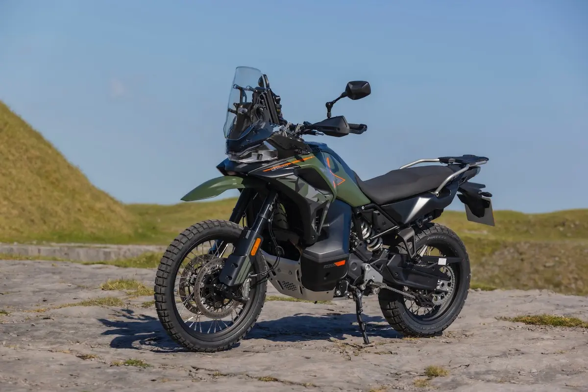 A green adventure motorbike, featured in a recent CFMOTO 1000MT-X review, stands on off-road tyres atop a rocky surface outdoors beneath a clear blue sky.
