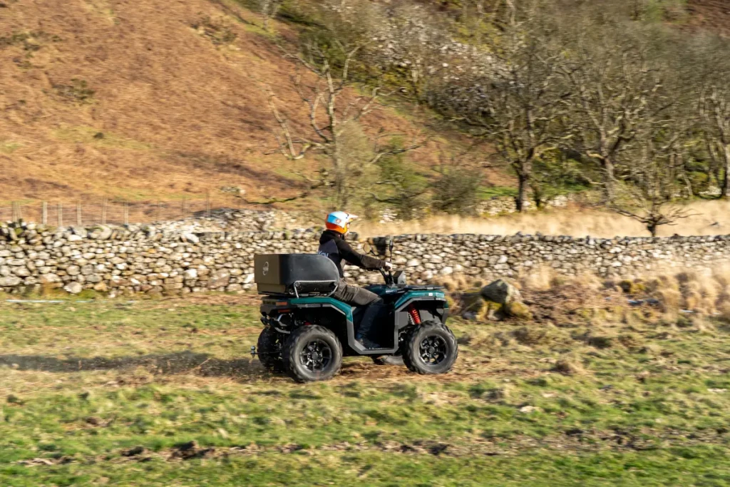 DSC01100 A person wearing a helmet rides a quad bike across a grassy field with a stone wall and leafless trees in the background.