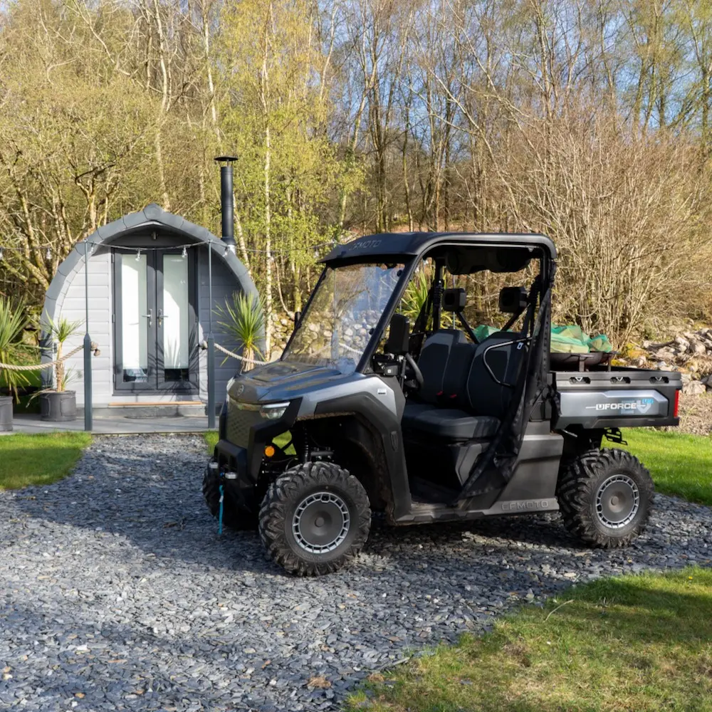 Holiday park utility vehicles A black holiday park utility vehicle is parked on a gravel drive in front of a small, modern pod-shaped cabin surrounded by trees and grass.