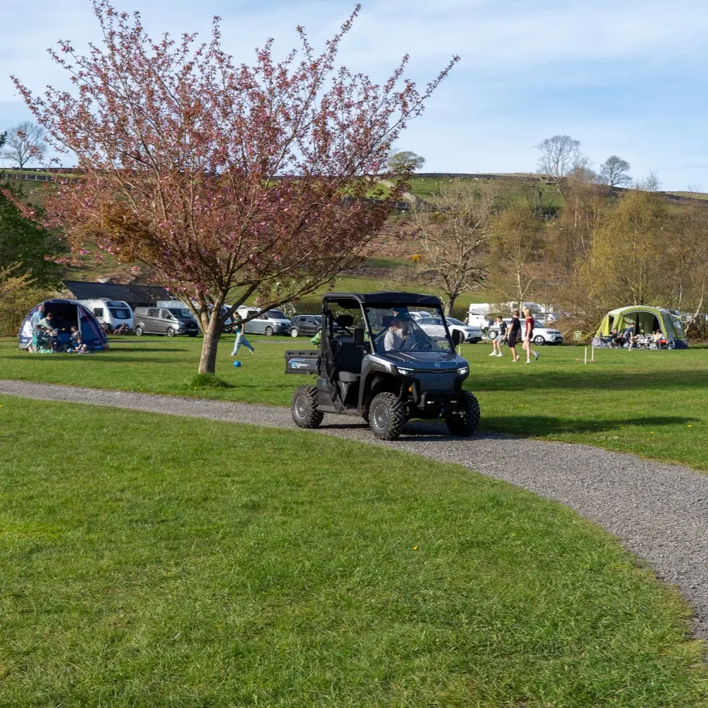 Holiday park utility vehicles A black holiday park utility vehicle drives on a gravel path through a grassy campsite with tents, people, and parked cars in the background.