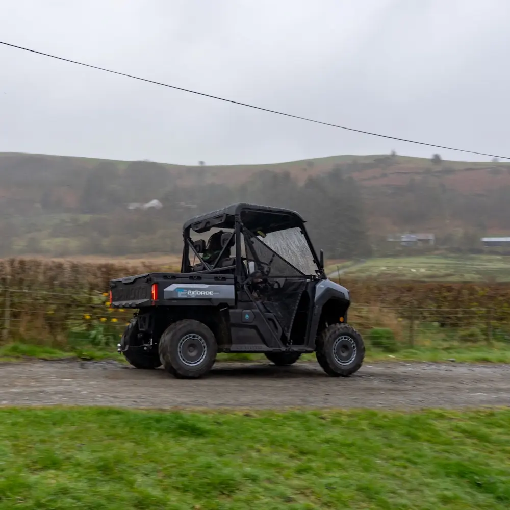 Holiday park utility vehicles A black holiday park utility vehicle driving on a gravel track with grassy fields and hills in the background under a cloudy sky.