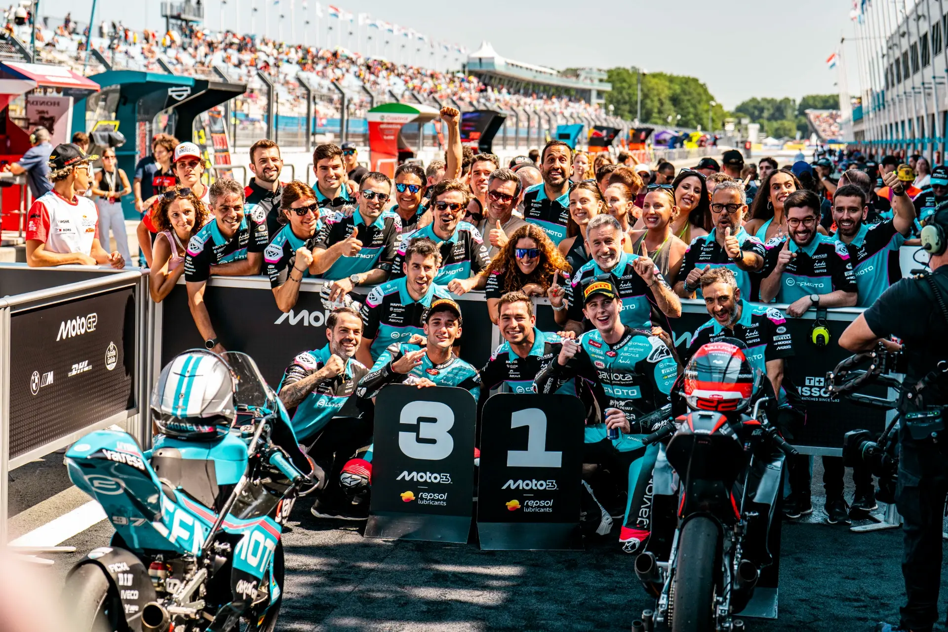 A CFMOTO motorcycle racing team poses for a group photo on the track, celebrating first and third place finishes with their bikes and podium boards at the Dutch MotoGP Grand Prix.