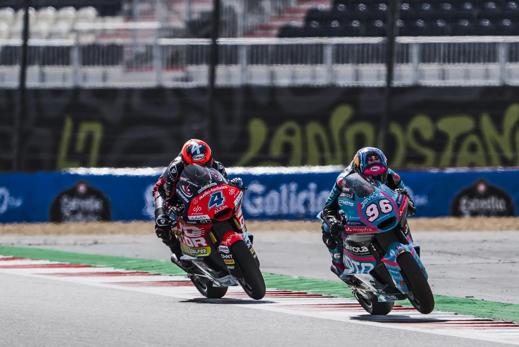 Two motorcycle racers in full kit compete on a racetrack at the British Grand Prix, leaning into a bend with blurred stands and fencing in the background, showcasing the high-speed action of racing.