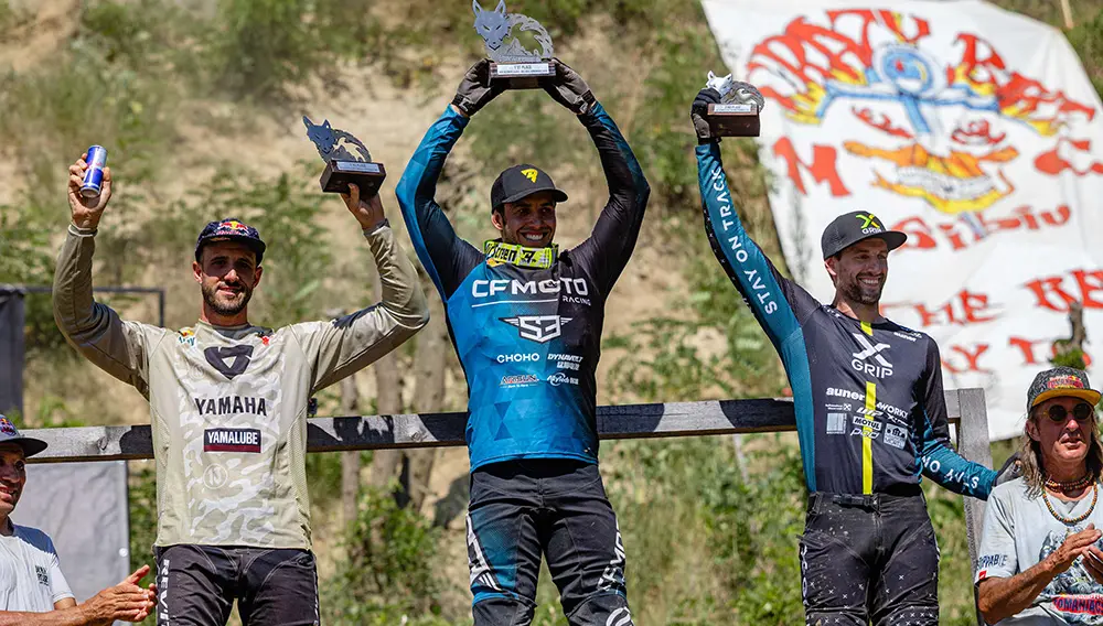 Three motocross riders stand on a podium holding trophies, celebrating their victory at the Red Bull Romaniacs event. A large CFMOTO banner and enthusiastic spectators are visible in the background.
